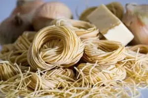 Uncooked nests of Capellini pasta, also known as angel hair, displayed with whole garlic bulbs in the background.