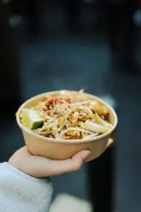 A hand holding a takeaway bowl of thick noodles topped with green onions and lime, demonstrating a modern street-food style serving of rustic Bigoli pasta.