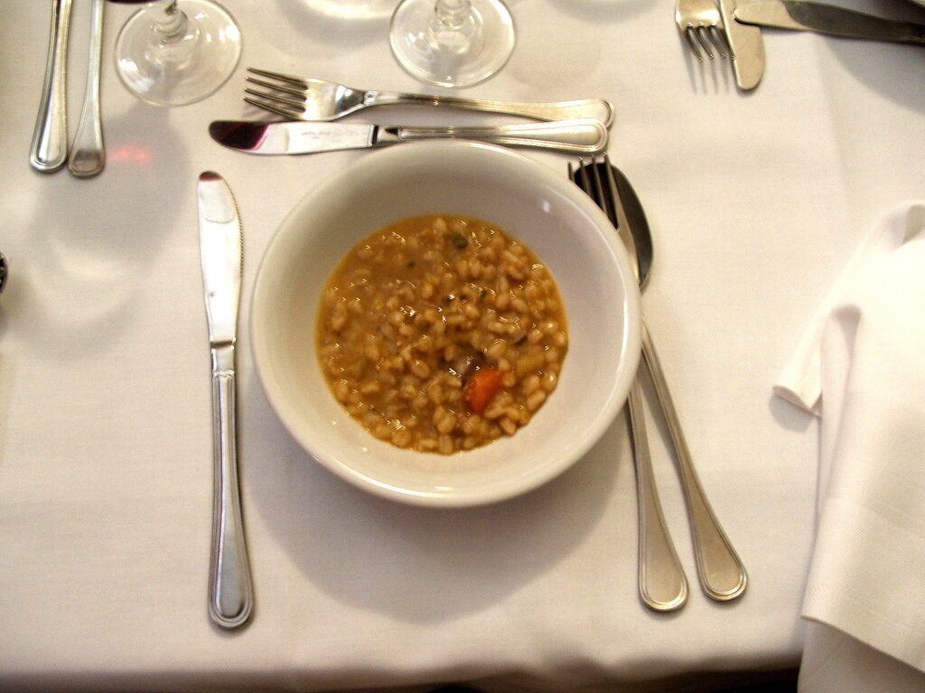 A rustic bowl of thick Italian farro pasta soup served on a white tablecloth with a fork and spoon.