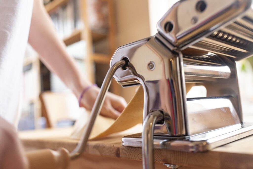 A close-up shot of fresh pasta dough being rolled through a shiny, stainless-steel manual hand-crank pasta machine one of the 7 essential tools for homemade pasta