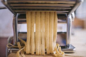 A close-up shot of fresh pasta dough being rolled through a shiny, stainless-steel manual hand-crank pasta machine, one of the 7 essential tools for homemade pasta.